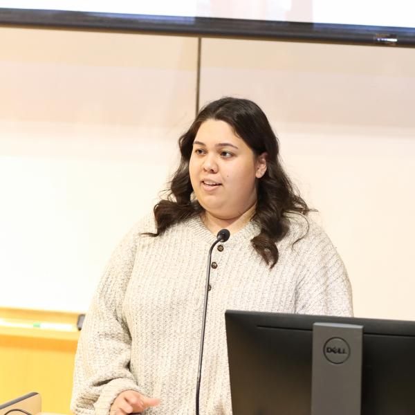 A student wearing a white sweater stands in front of a microphone giving their presentation.