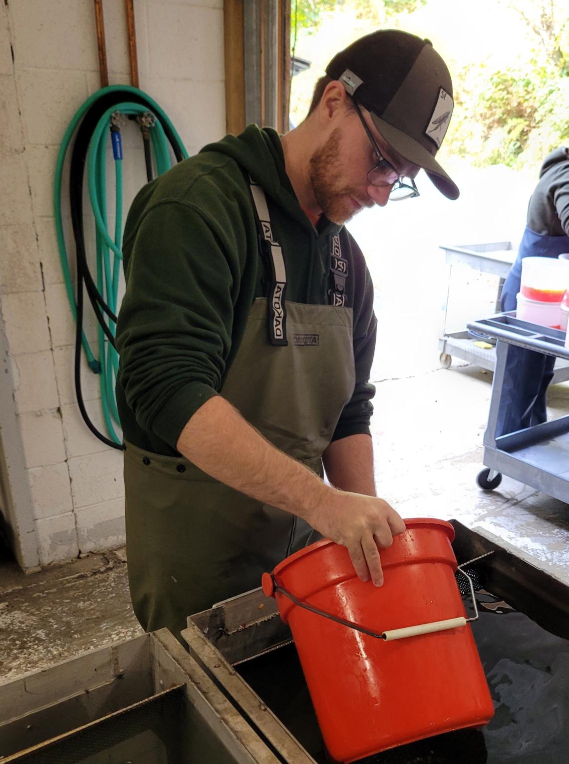 A young man in hip waders is holding an orange bucket over a tray