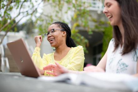 Two students sit at a table outdoors, one has a laptop open and the other has a textbook open. They are laughing together