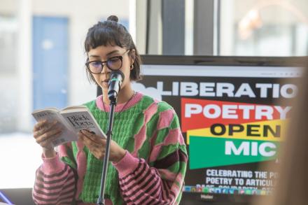 A woman wearing glasses and a pink and green sweater reads from a poetry book at a microphone