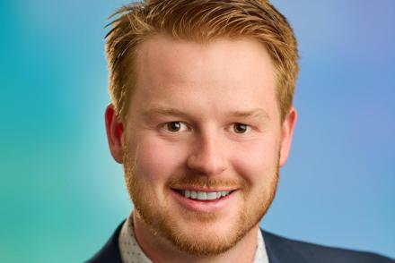 Mitch Turko wearing a suit and tie, smiling at the camera with a blue and green background.