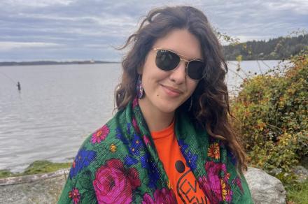 Alyona stands near a lake wearing a green shawl with pink, blue and yellow flowers over an Every Child Matters shirt.