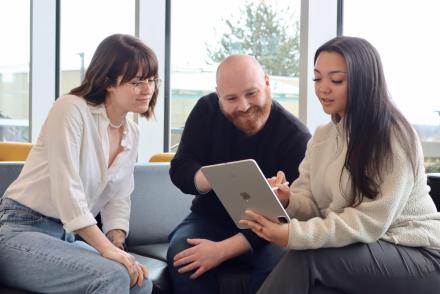 A woman, a man and another woman sitting on a chesterfield looking at a laptop together.