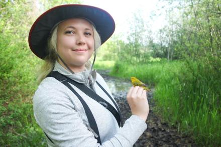 Chelsey Watts stands amongst bushes and trees holding a bird.