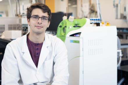 Joseph Monaghan sits in a VIU Chemistry lab, wearing a white lab coat.