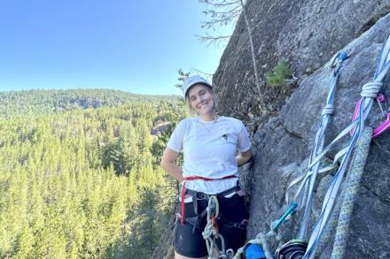 Sydney Solland stands on a ledge next to a rock face with climbing gear in front of her