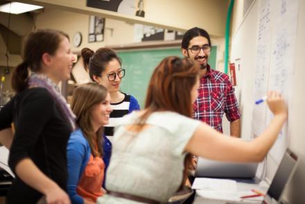 A group of students gathers around a student writing on a chalkboard