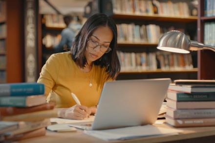 A woman sits at a desk in a library with a laptop open surrounded by books