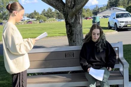 Two actors rehearse with scripts in their hand near a bench in a park.