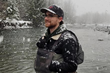 A young man clad in hipwaders is standing in a river with snowflakes falling around him